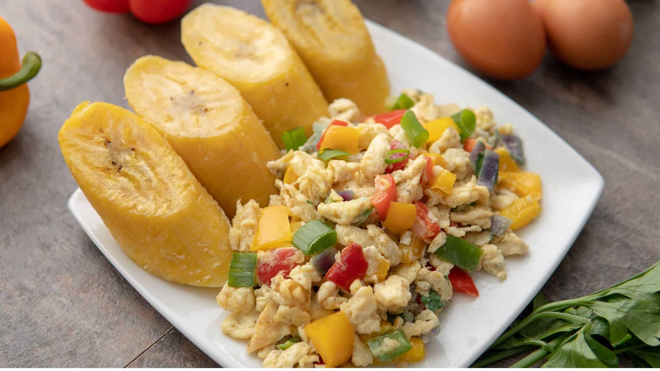 Fresh fried plantains served with scrambled eggs and colorful diced vegetables on a white plate, surrounded by fresh tomatoes and herbs, perfect for a nutritious meal.